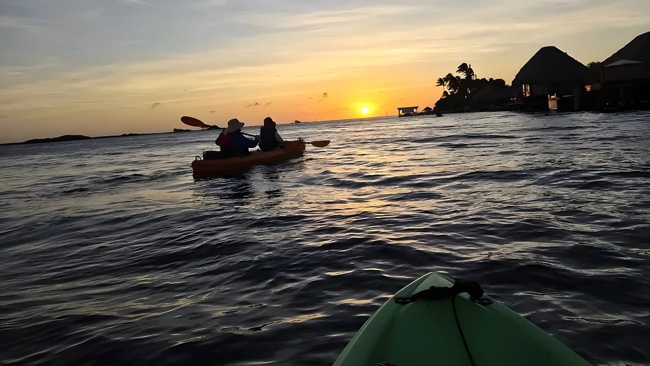 The view over the bow of a kayak at sunset off Aruba's south coast, a tandem kayak ahead and the sun going down behind the palms