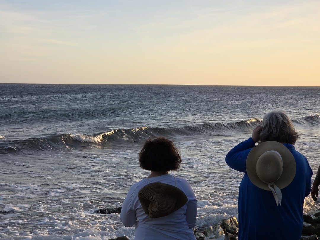 Two people in sun hats, seen from behind, watching a wave roll in at the rocky surf line under a warm evening sky