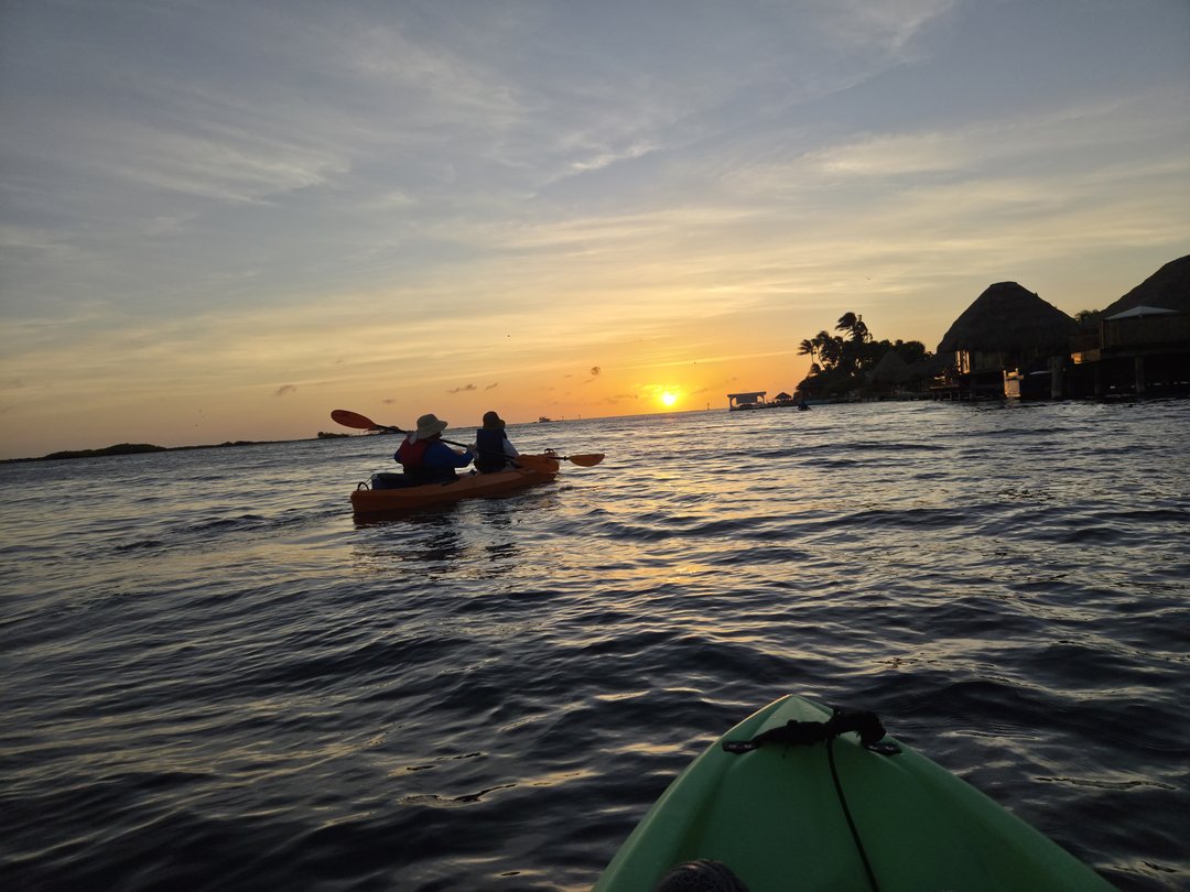 A double kayak ahead on flat water with the sun setting behind palm-roofed buildings, our own bow in the corner of the frame