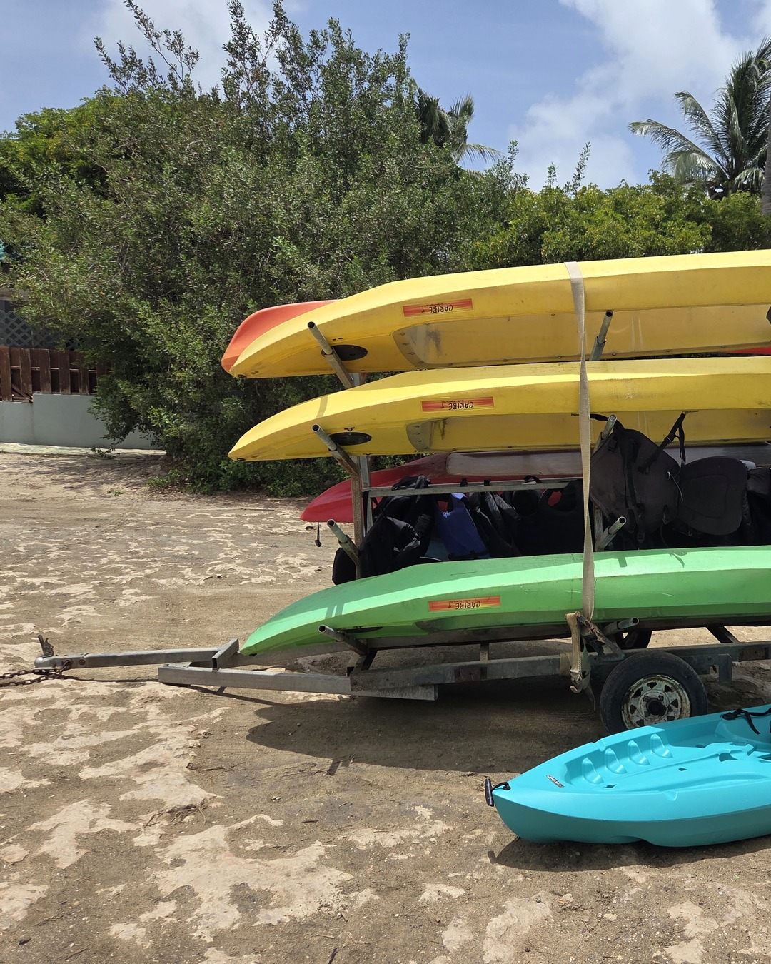 Yellow, red and green kayaks strapped to a trailer rack with life jackets hanging beneath, a turquoise kayak resting on the sand alongside