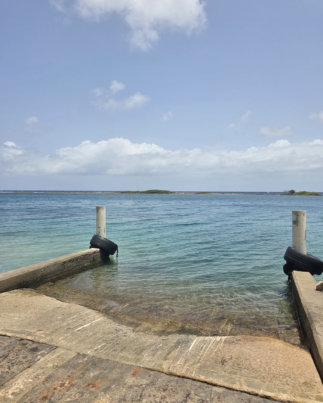A concrete launch ramp running into shallow turquoise water between two tire-capped mooring posts, a low green islet on the horizon